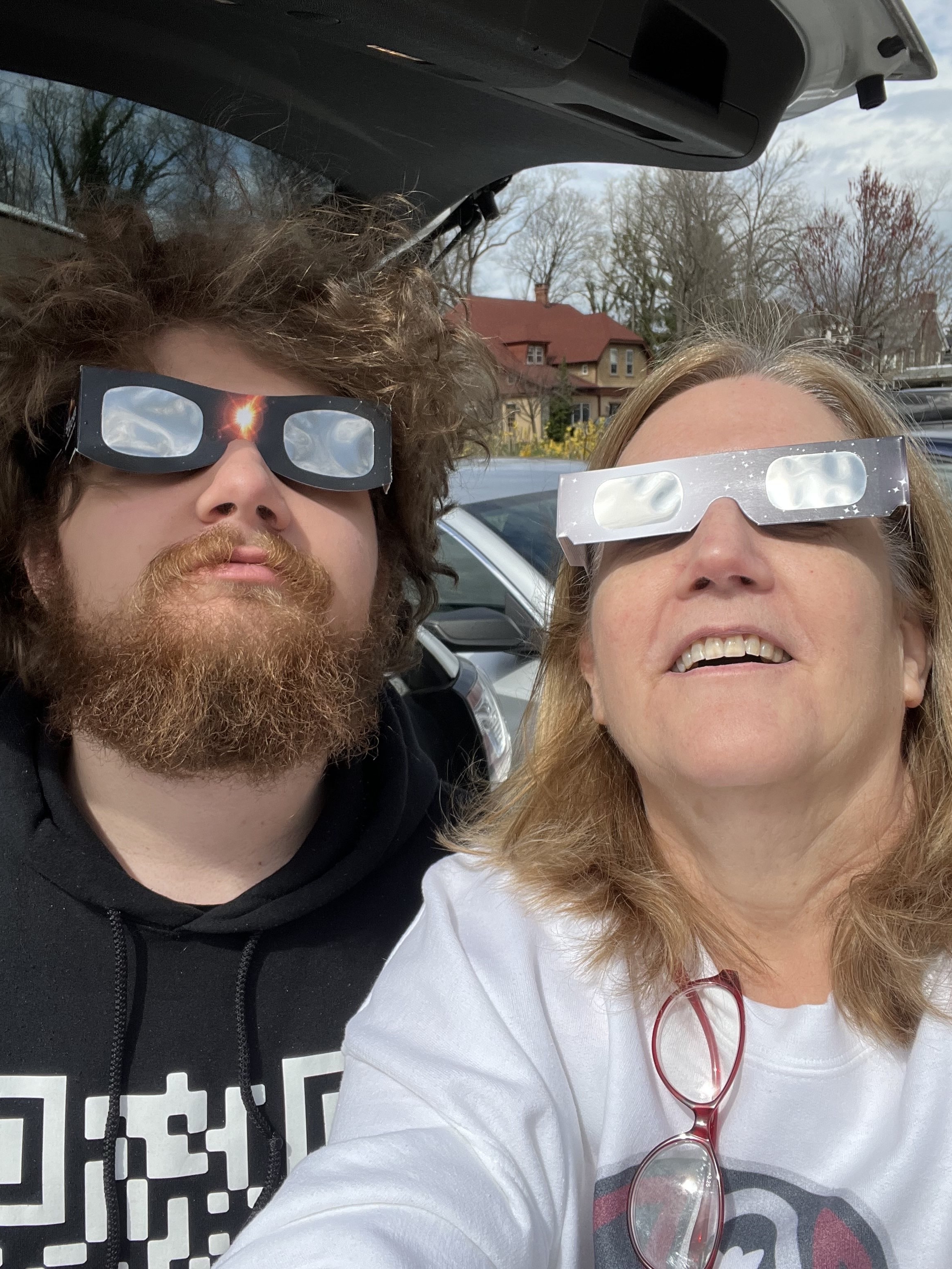 A rectangular photograph of a young white man and a middle-aged white woman wearing reflective solar eclipse glasses, looking up at the sky. The man has curly brown hair and a short, thick beard, and is wearing a black hoodie. The woman has long, blonde hair and is wearing a white shirt.